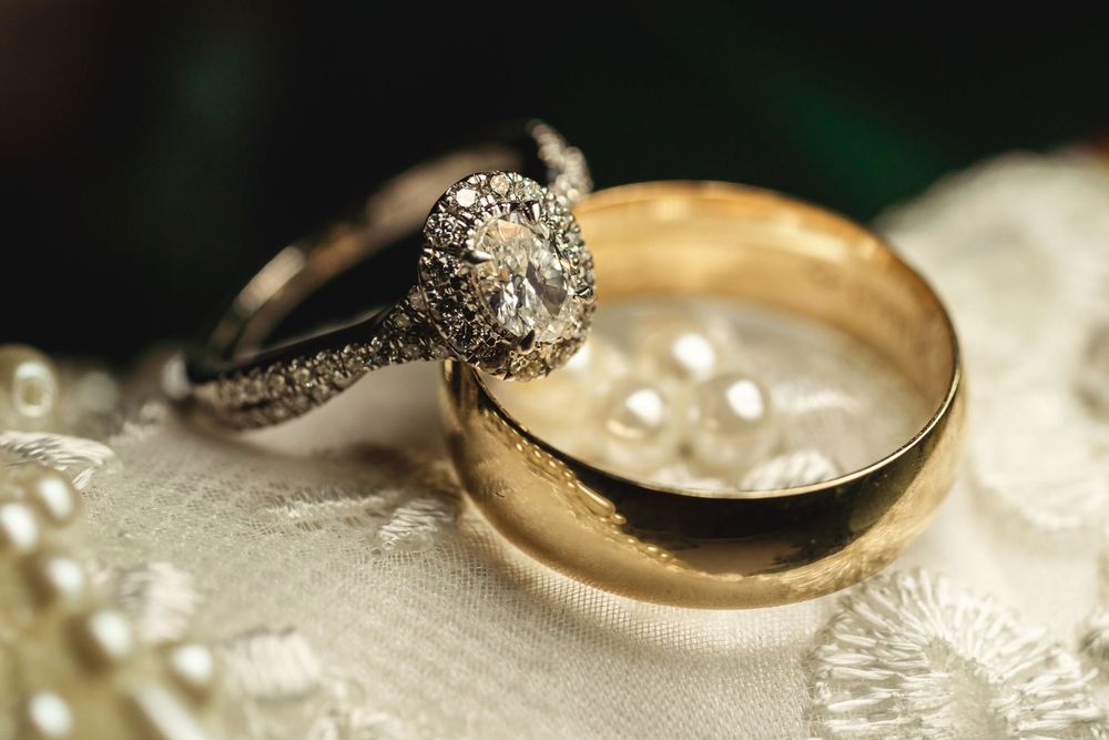 Diamond engagement ring and gold wedding band rest on delicate white lace fabric with scattered pearls in soft lighting.