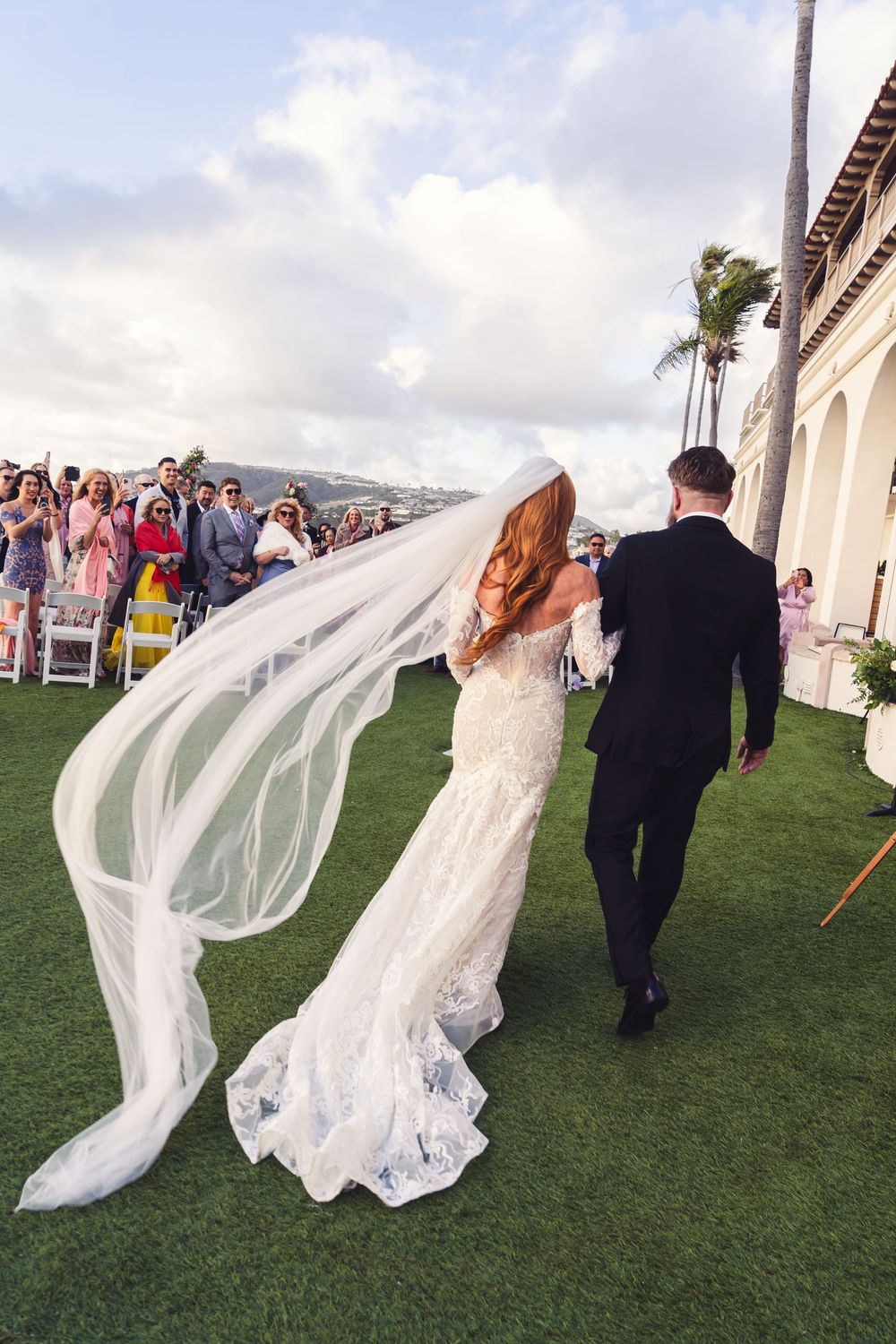 A long flowing veil catches the wind as a couple walks across an outdoor wedding at ritz carlton dana point with guests in the background.