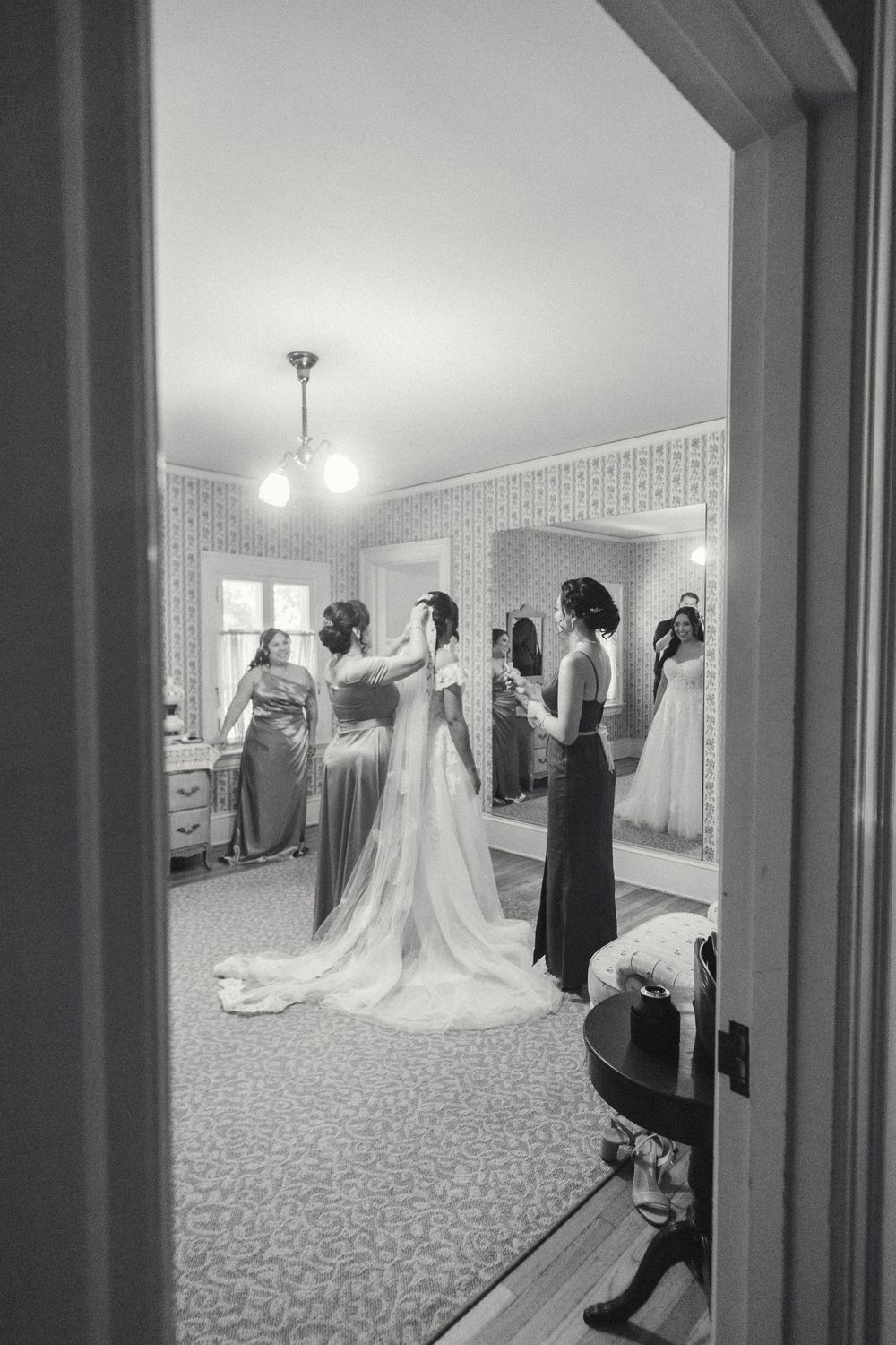 Black and white photograph showing a bridal party getting ready in a mirror at Lindley Scott House with a long white wedding gown.