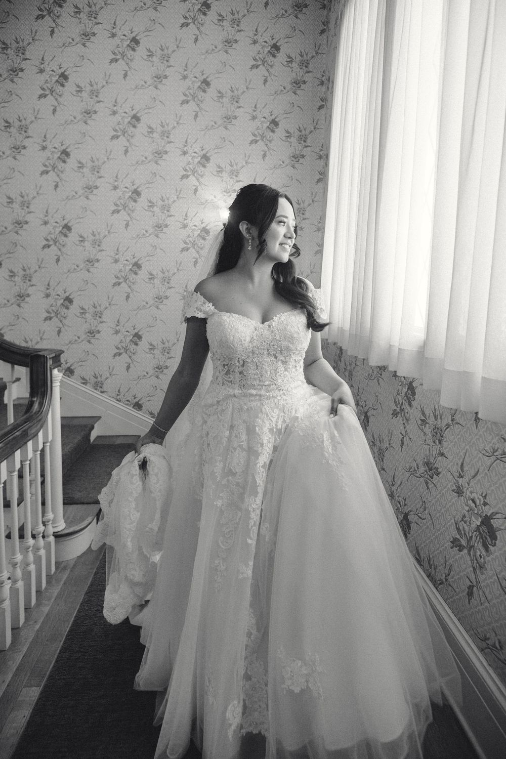 A bride in a beautiful off-shoulder lace ball gown stands elegantly by a window with floral wallpaper in black and white Lindley Scott House