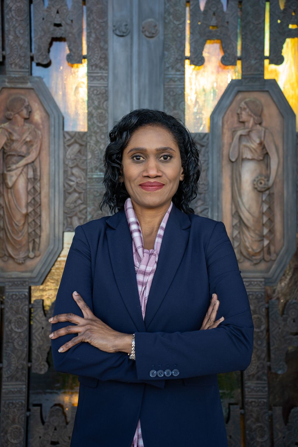 Professional portrait of a person in a navy blazer and pink striped shirt standing in front of an ornate carved wall.