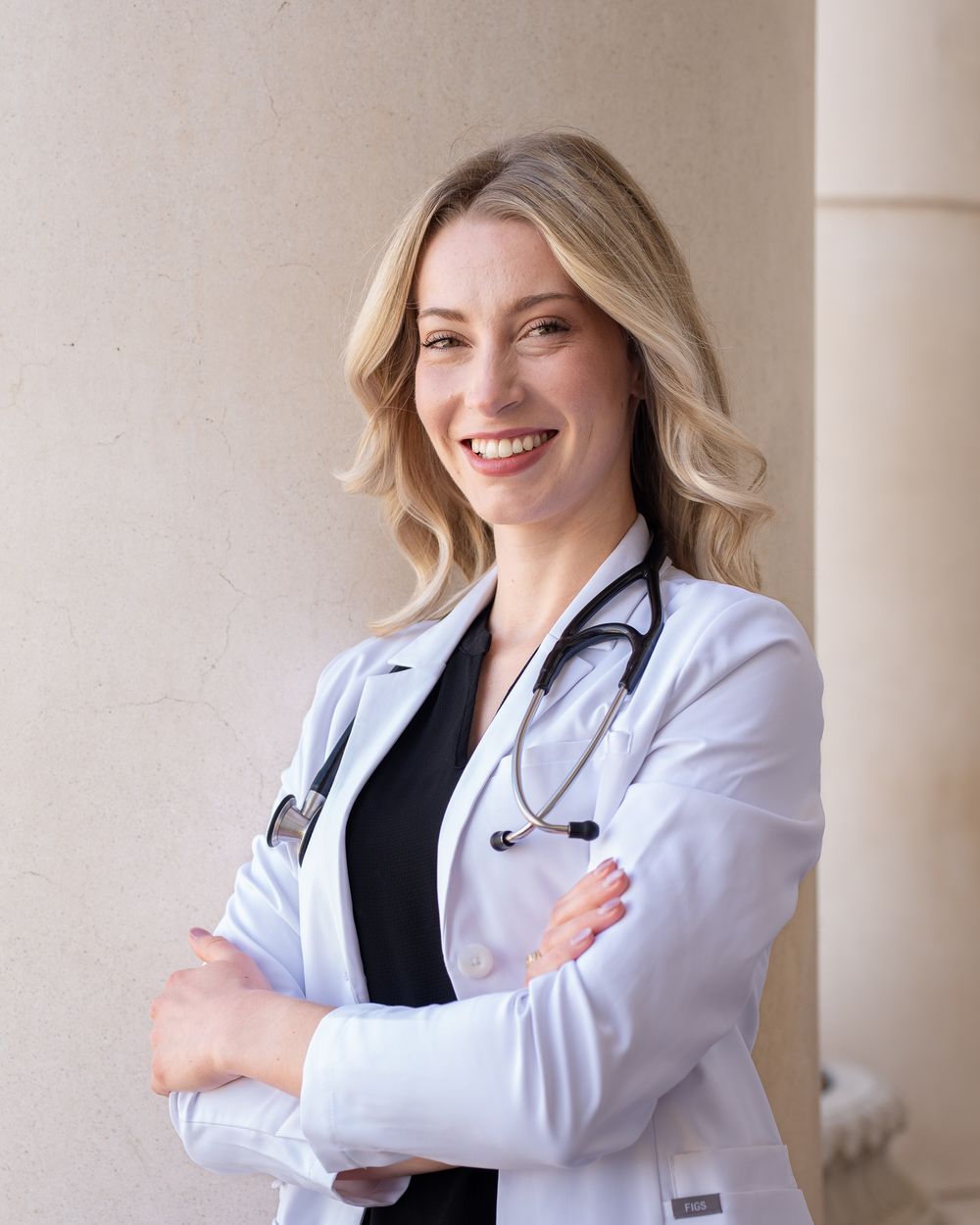 A medical professional in a white coat with stethoscope poses confidently with crossed arms against a neutral background at Teaxs A&M