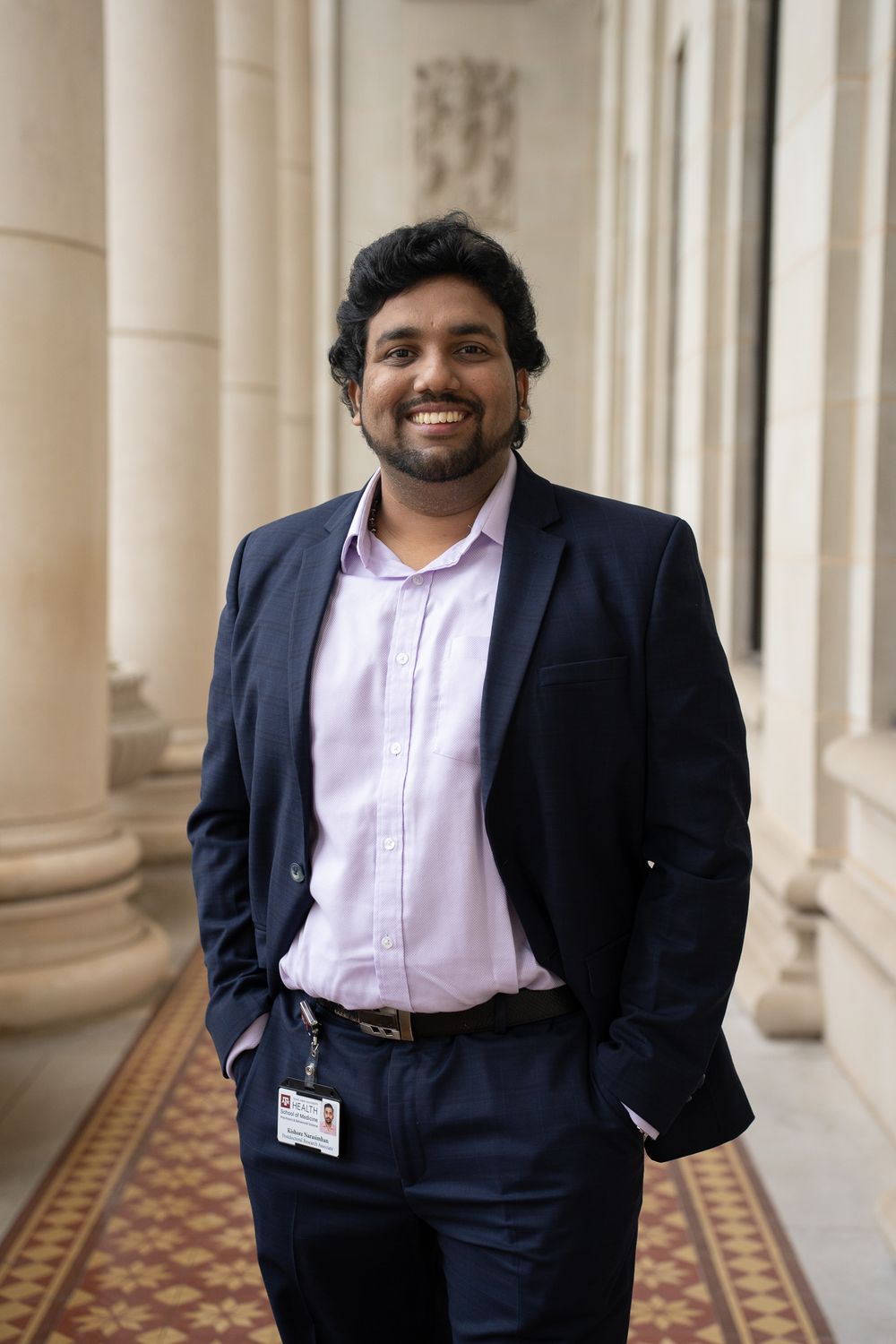 Professional in a navy suit and pink shirt standing in a columned hallway.