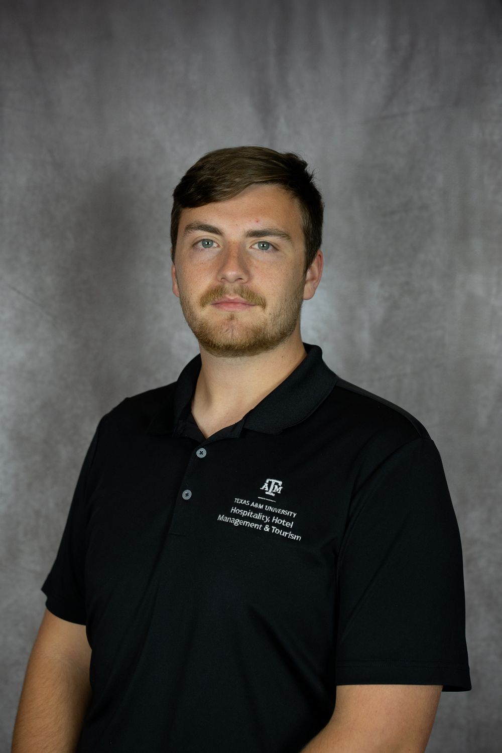 Professional headshot of someone wearing a black polo shirt with embroidered logo against grey background.
