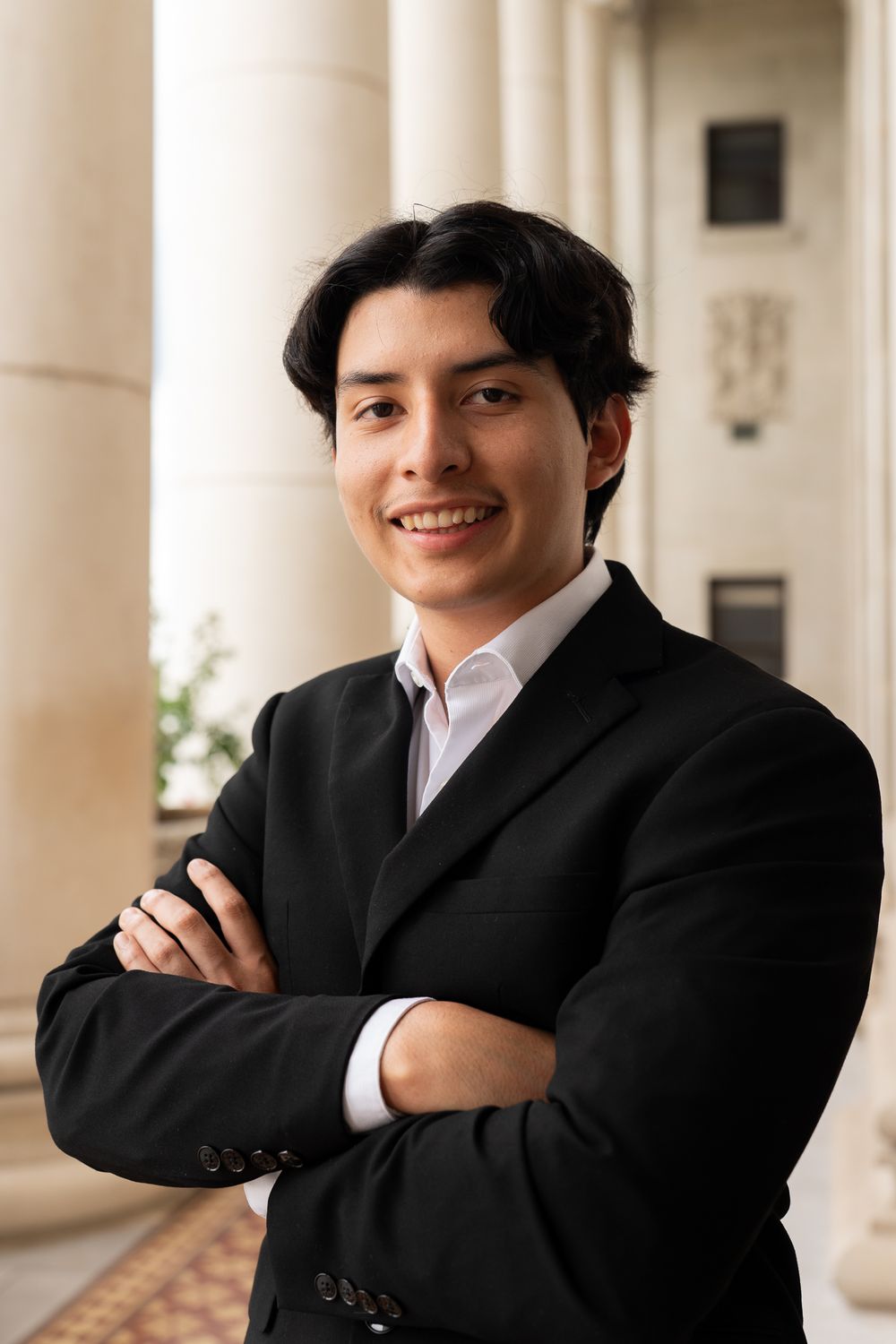 Professional headshot of a young professional in a black suit and white shirt standing with crossed arms in an office hallway.