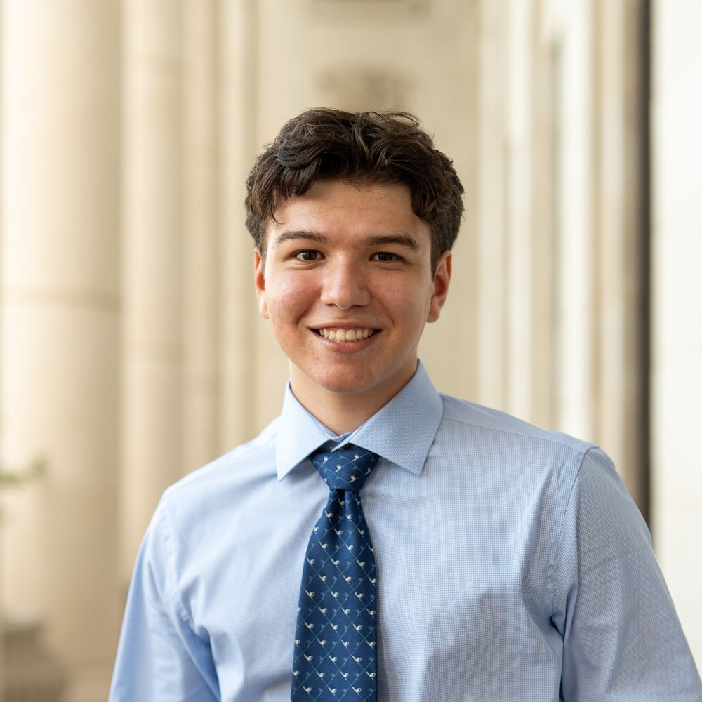 Professional portrait of someone wearing a light blue dress shirt and blue patterned tie smiling against a cream background.