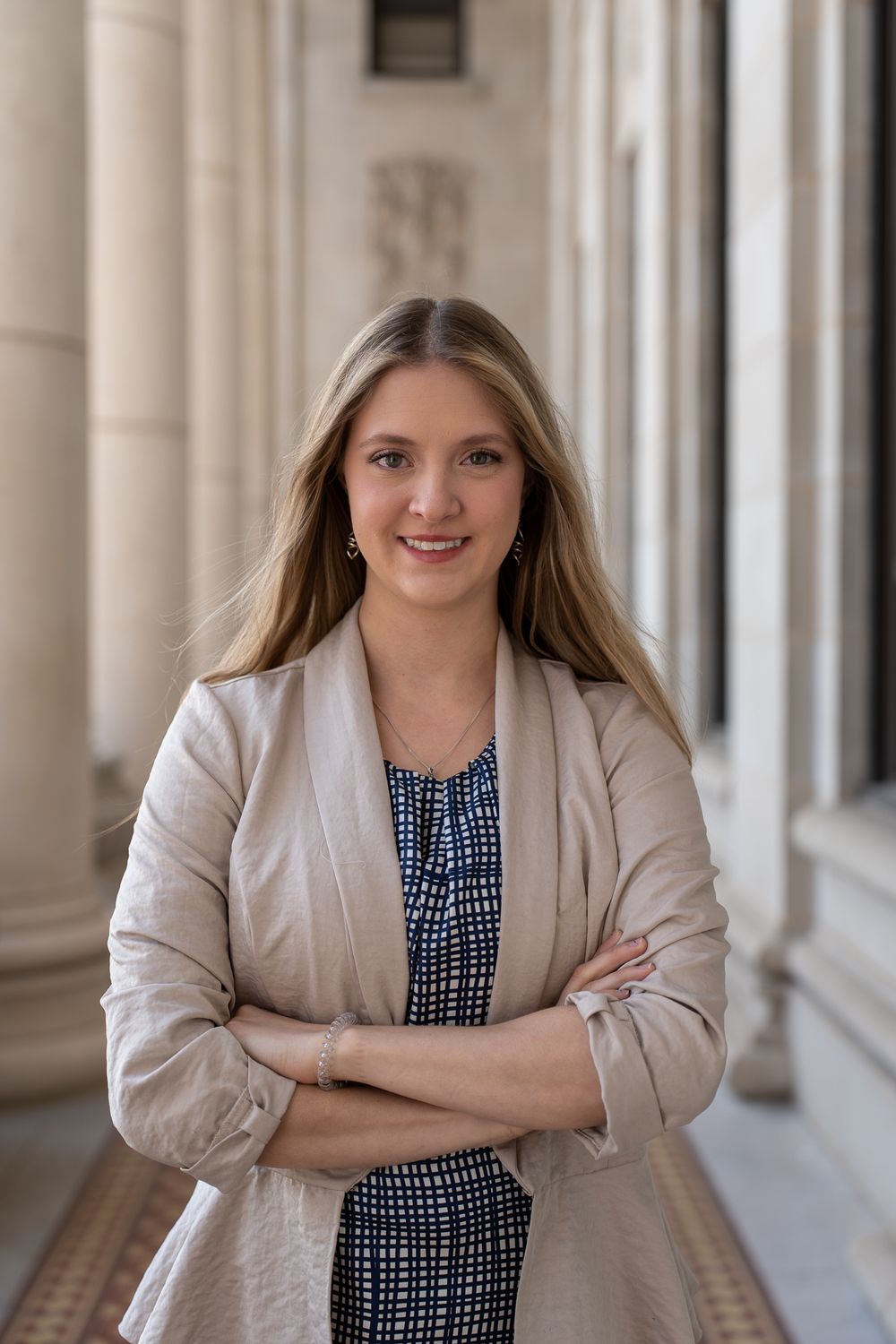 Professional portrait of someone in a beige blazer and patterned dress standing confidently in a columned hallway.