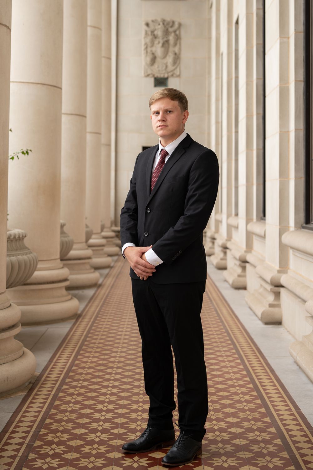 Professional in a dark suit and red tie stands in an elegant hallway with classical columns and patterned floor tiles.