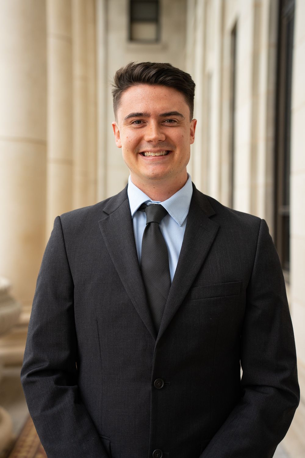 Professional headshot of someone wearing a dark suit and tie standing in front of a light-colored columned background.