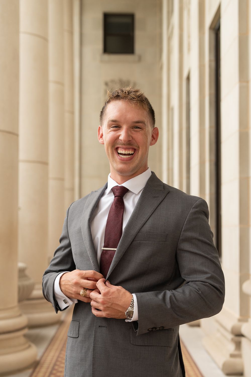 Professional wearing a gray suit and burgundy tie smiles warmly while standing between classic columns in formal setting.