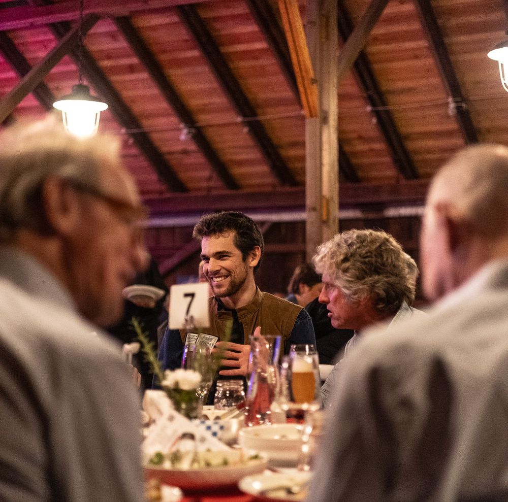 People gathered around a rustic wooden dining table for dinner with drinks and warm lighting overhead.