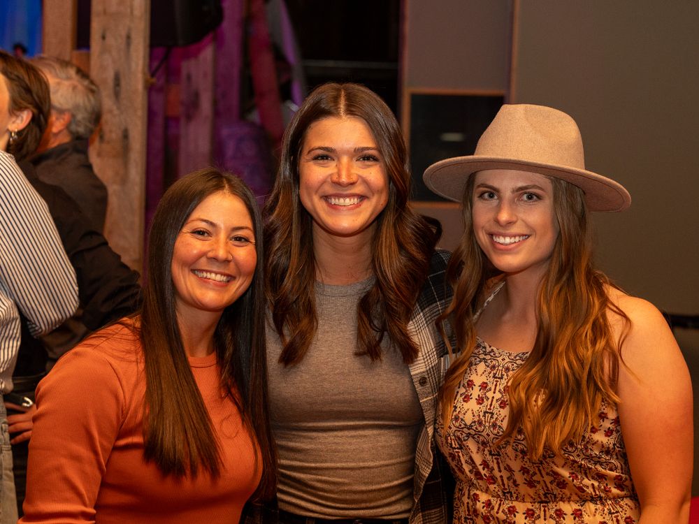 Three friends smile together at a social gathering, with one wearing a tan hat and another in an orange sweater.