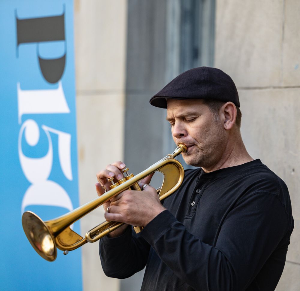 A musician in black plays a trumpet outdoors against a blue background.