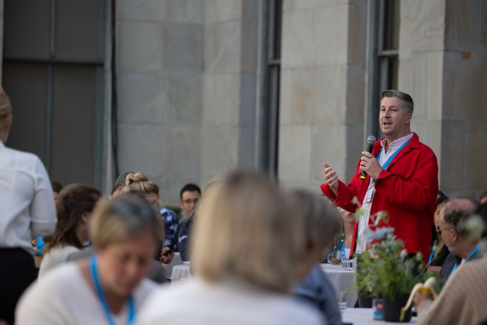 A speaker in a red jacket addresses an outdoor audience.