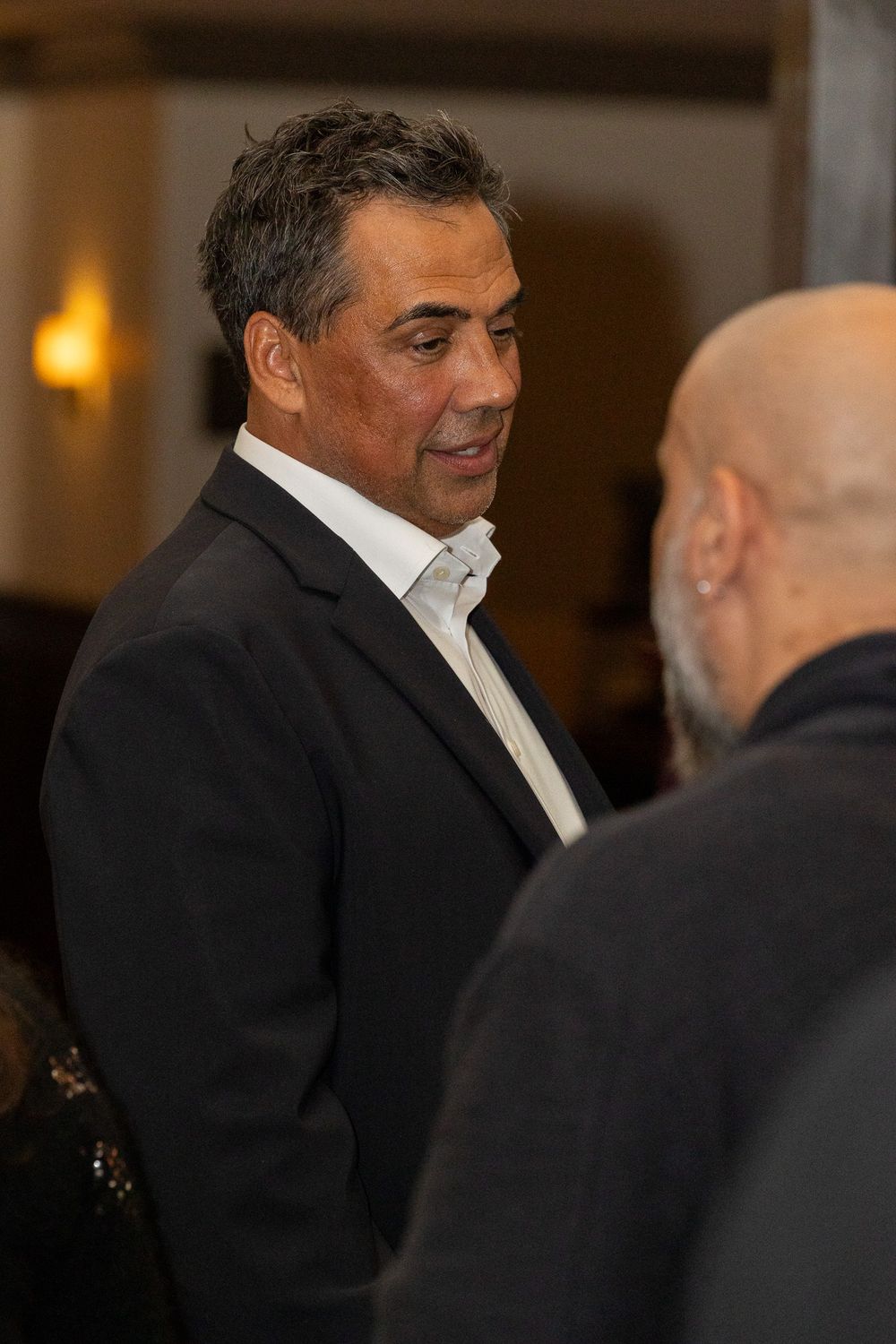 Two men in formal attire engaged in conversation at an indoor event with warm lighting in the background.