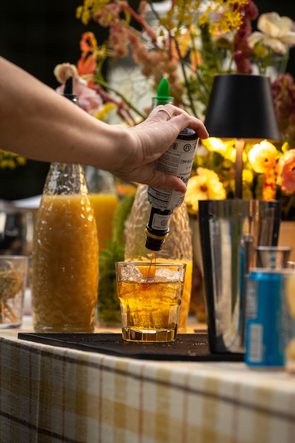 Bartender pouring a craft cocktail with colorful floral arrangements and bottles in background.