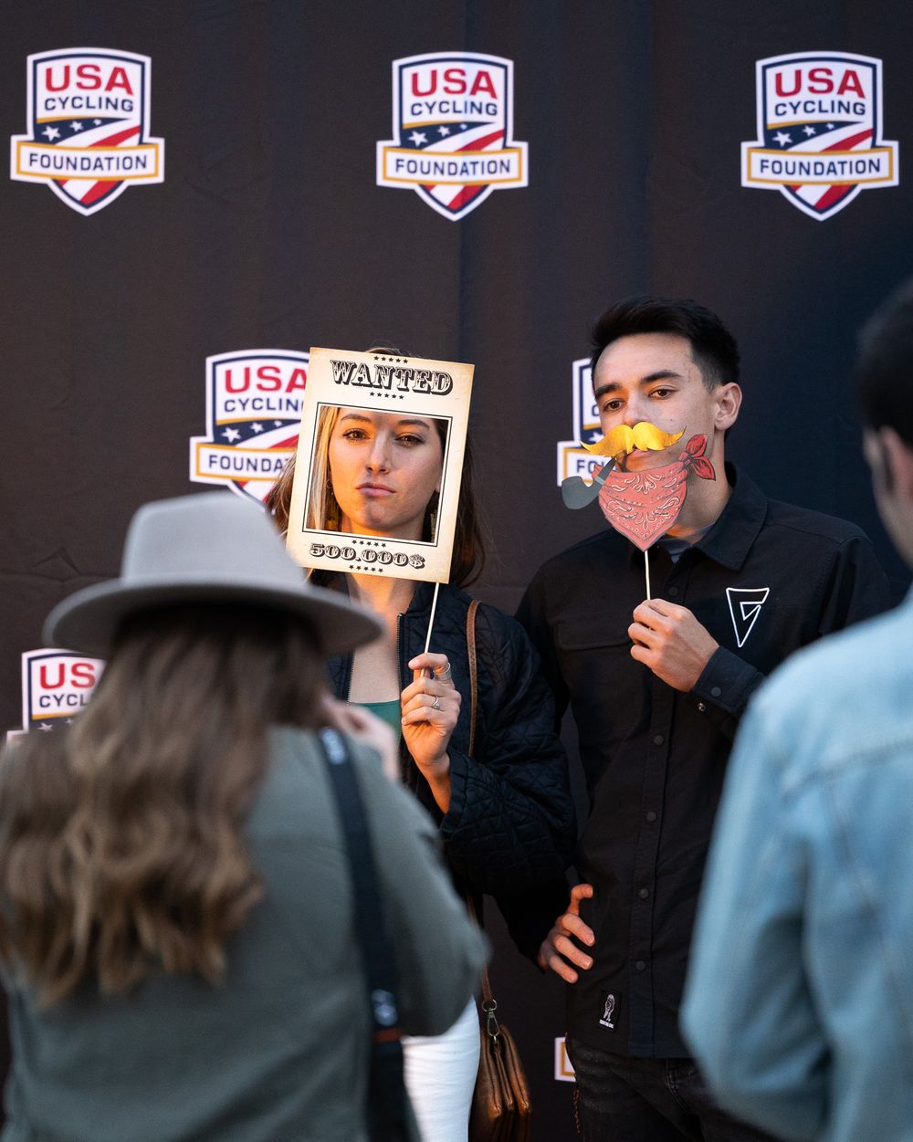 People pose with signs and decorative props at a USA event with multiple branded banners in the background.