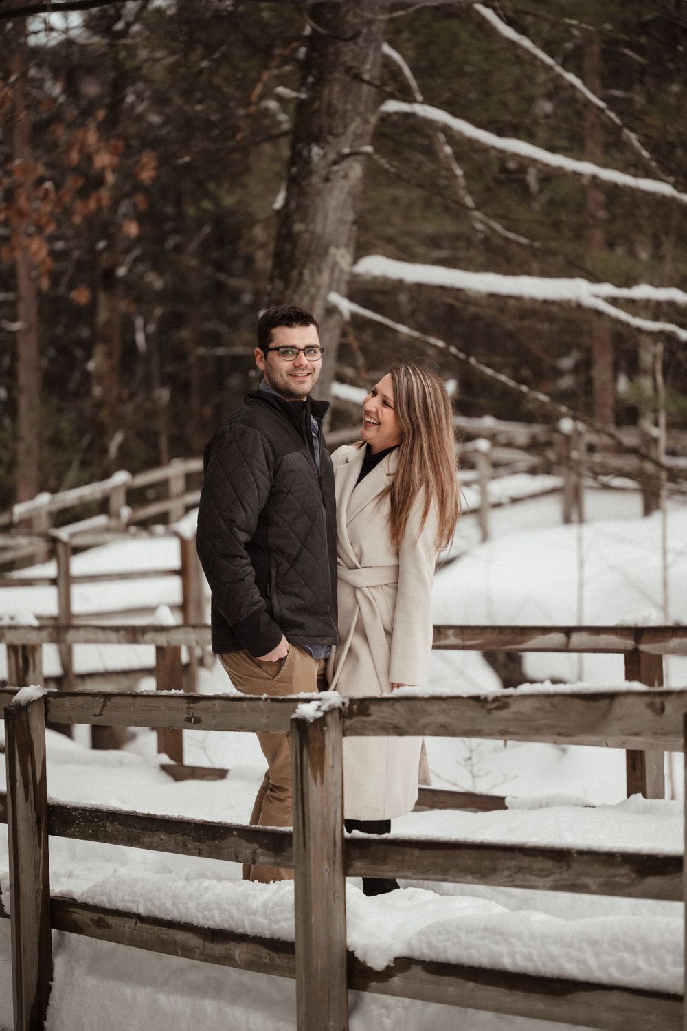Winter engagement portrait of couple standing on wooden fence in snowy forest setting.