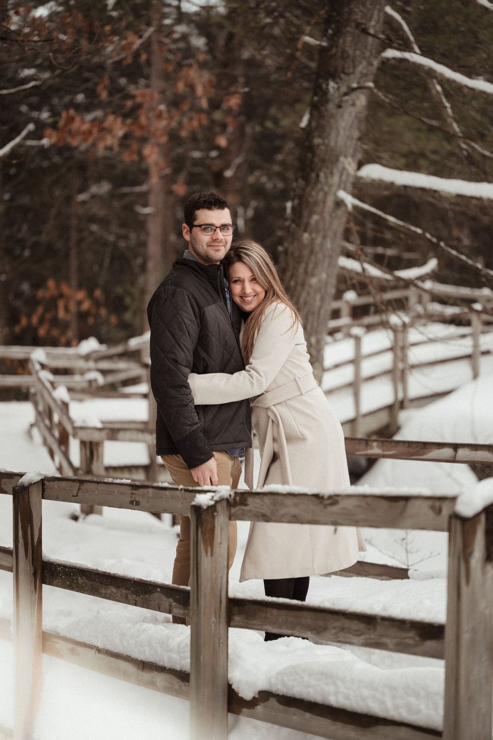 A couple embraces on a wooden fence walkway in a snowy winter forest setting.