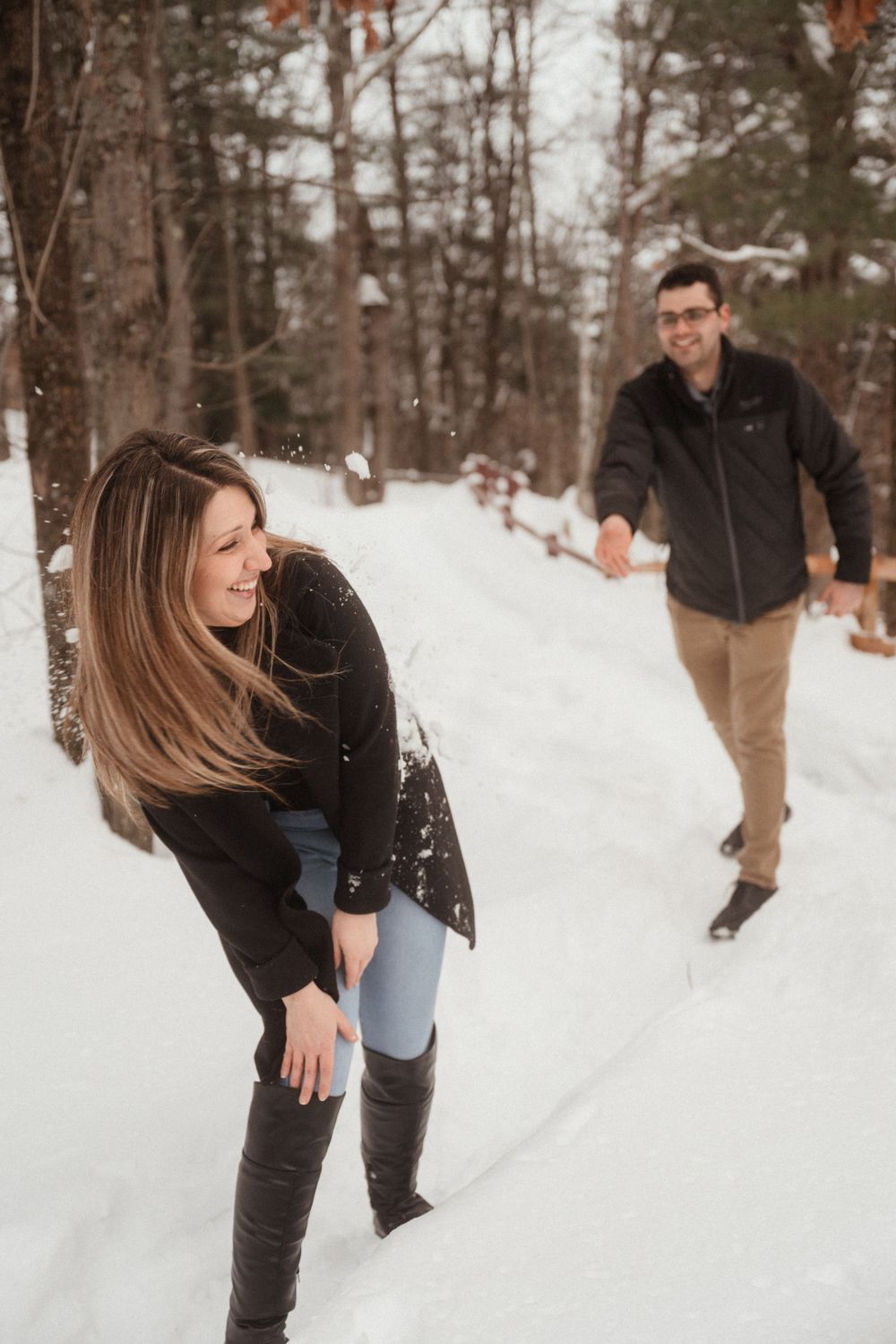 Two people playing and laughing in the snow along a winter forest path.