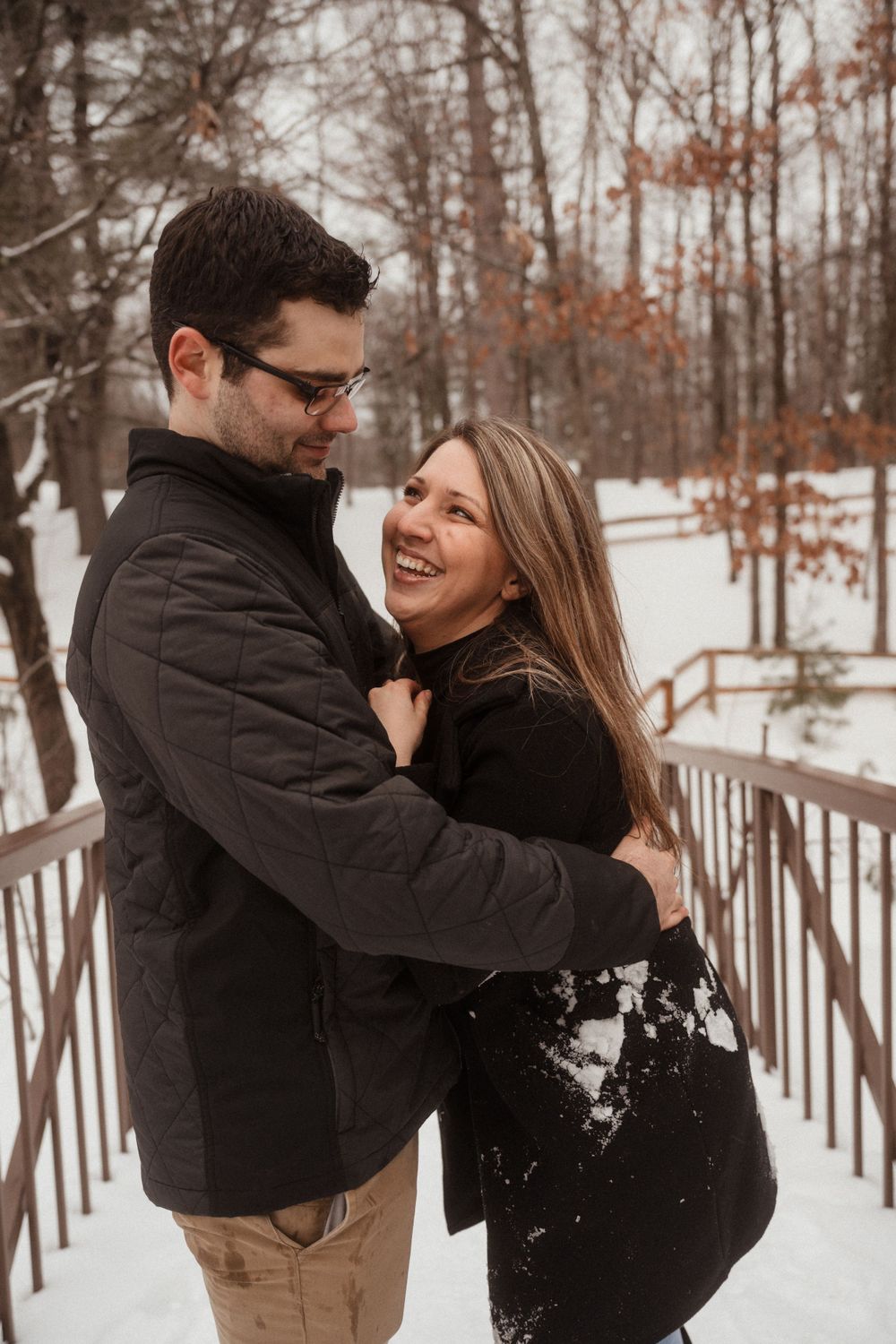 Couple embraces and laughs together on a snowy bridge in winter woods.