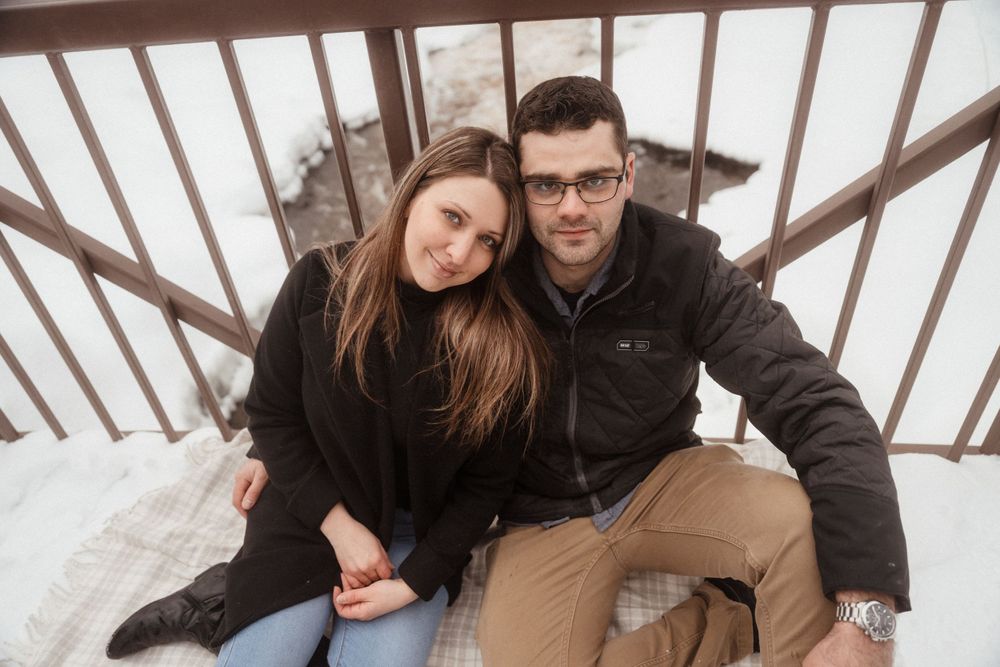 Couple in winter coats cuddling on wooden bridge railing.