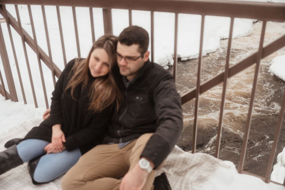 Couple sits close together on a snowy wooden bridge.
