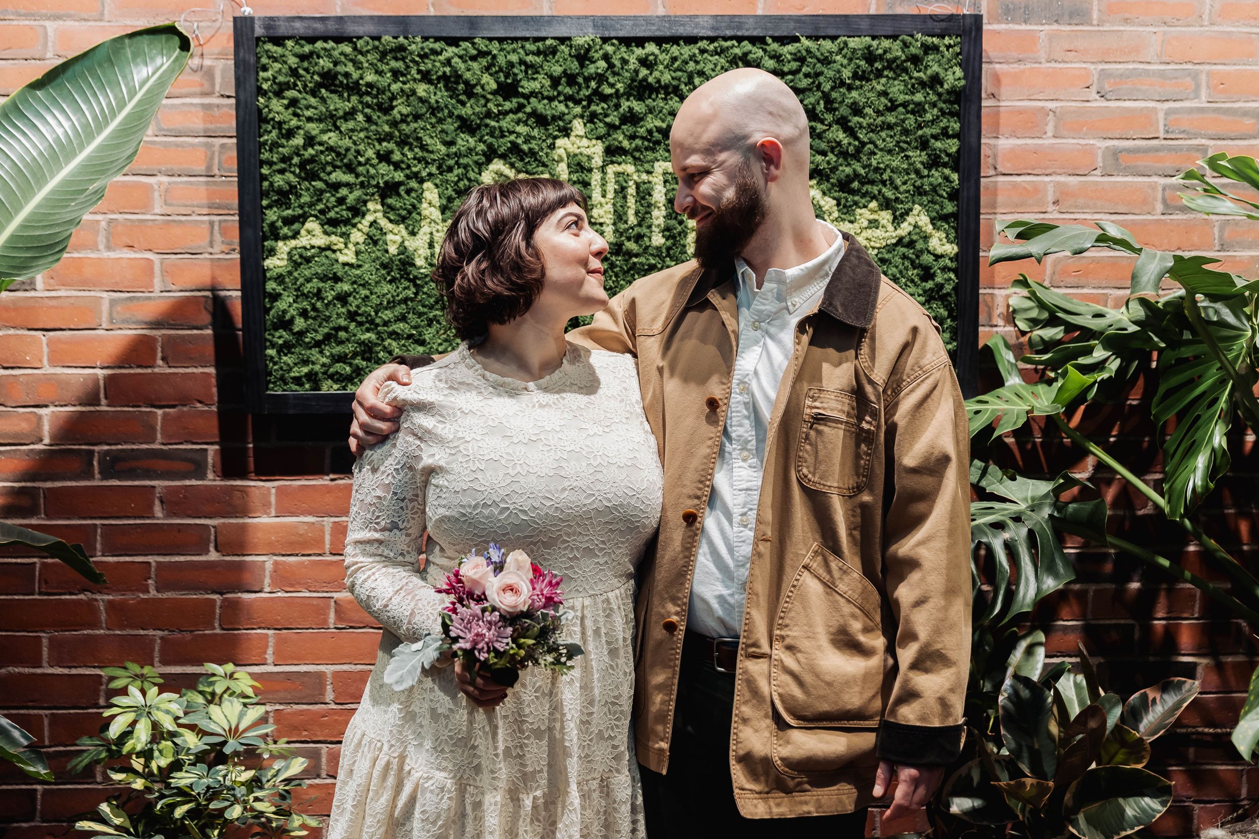 A couple poses for wedding photo against green hedge wall and brick background.