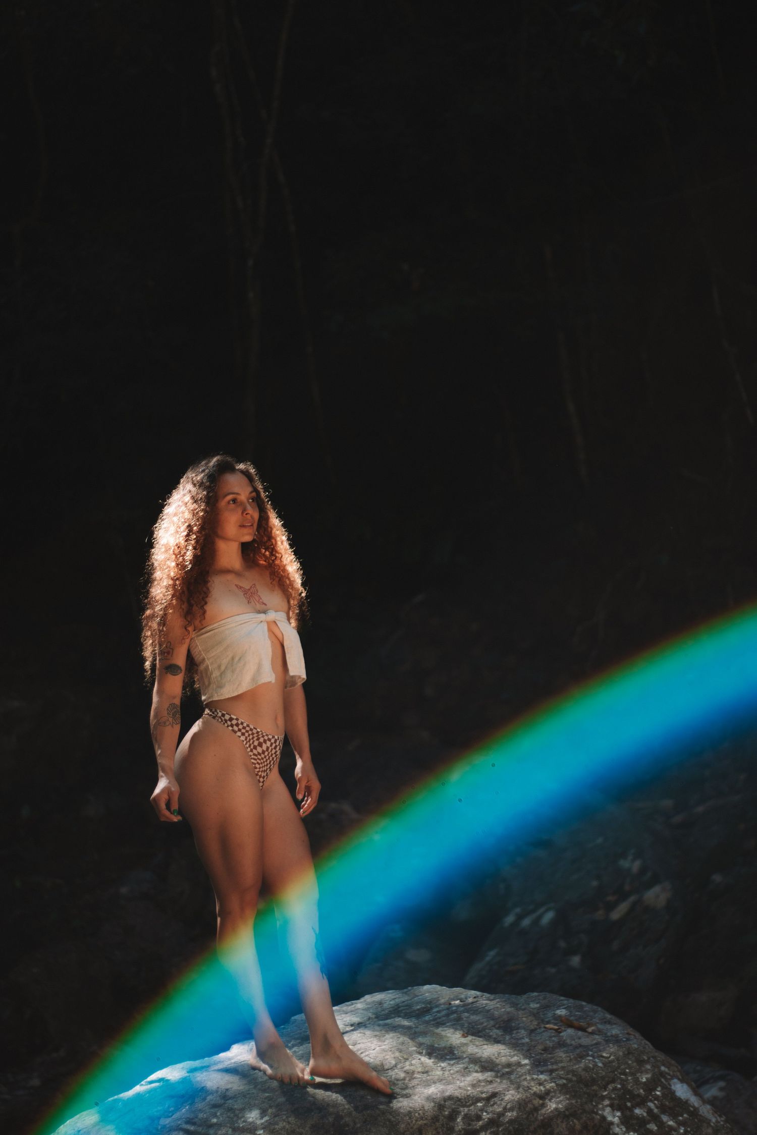 Model with curly hair standing on rock with natural rainbow across