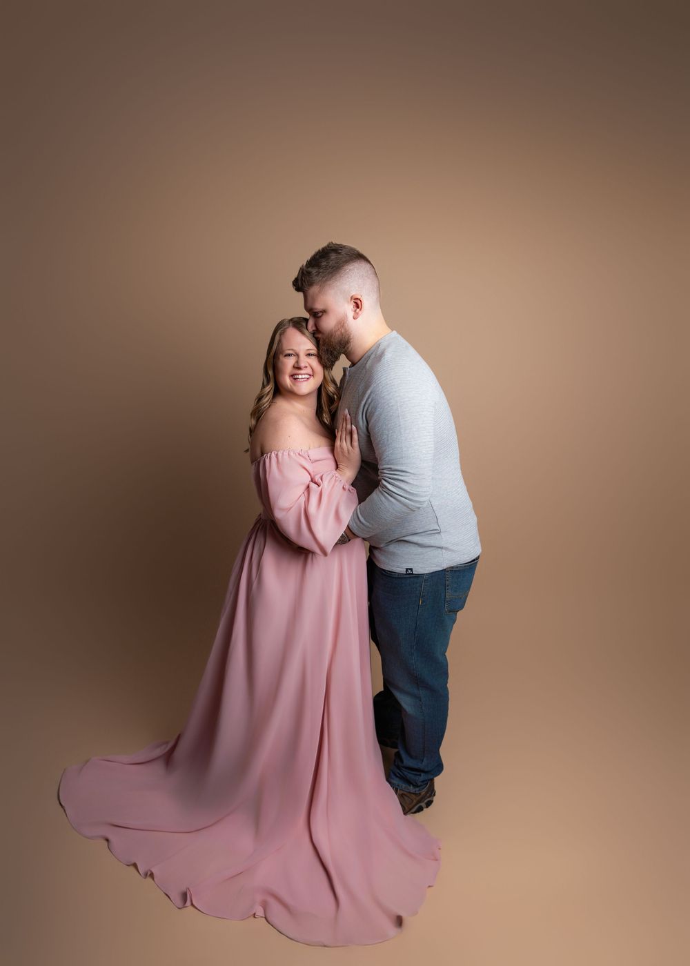 A romantic maternity studio portrait featuring a couple embracing, with her in a flowing pink gown and him in casual attire.