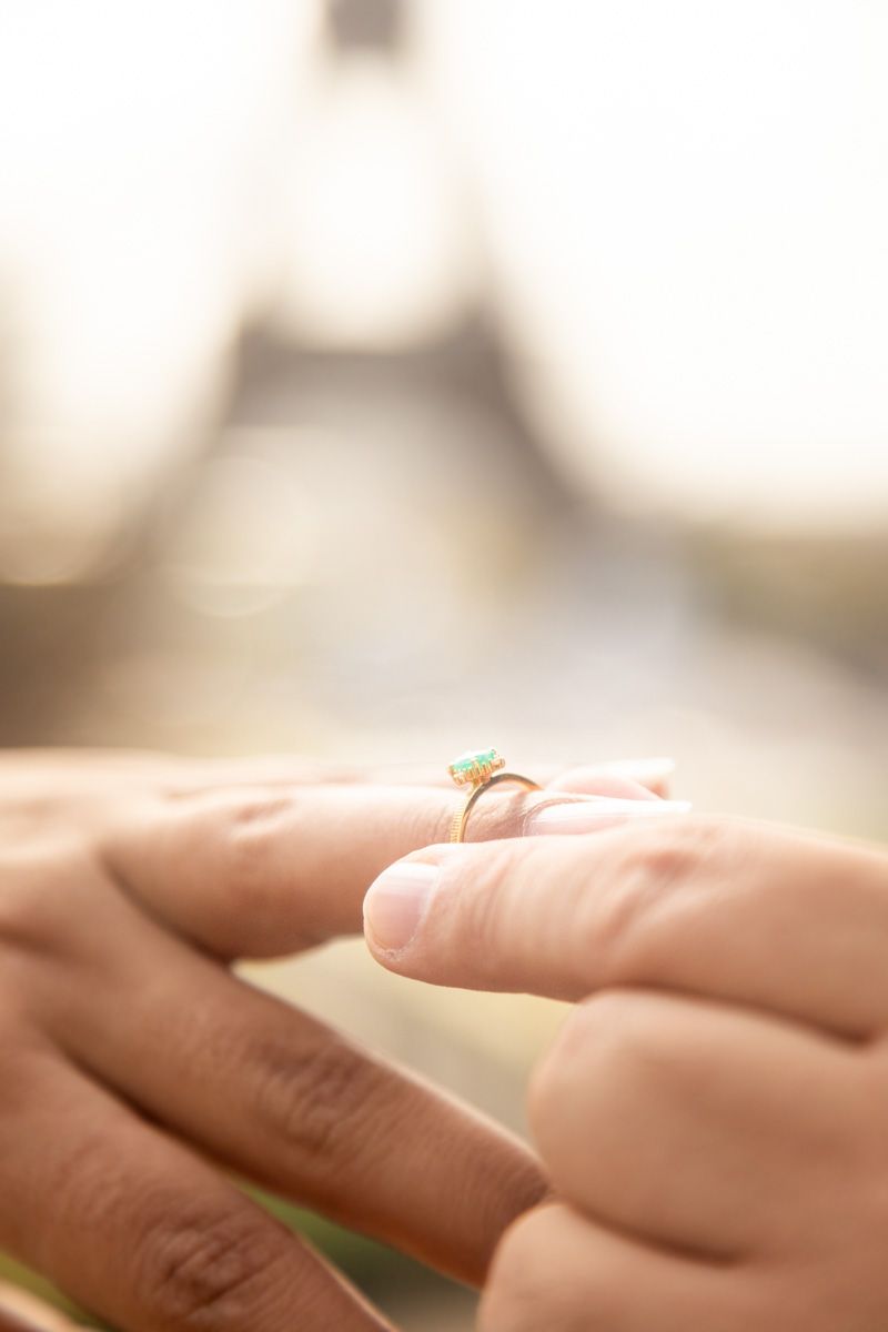 Close-up of hands holding an engagement ring with a blurred Eiffel Tower in the background.