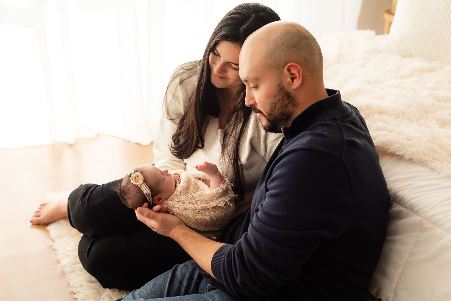 Lifestyle newborn portrait of mom and dad sitting on the floor next to a window holding and staring at their newborn baby girl.