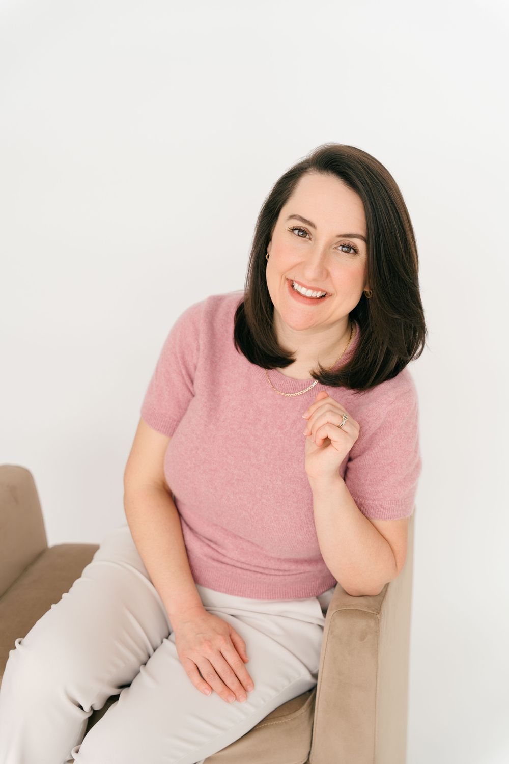 A woman in a pink t-shirt and light colored pants sits casually and smiles while posing in a neutral studio setting.