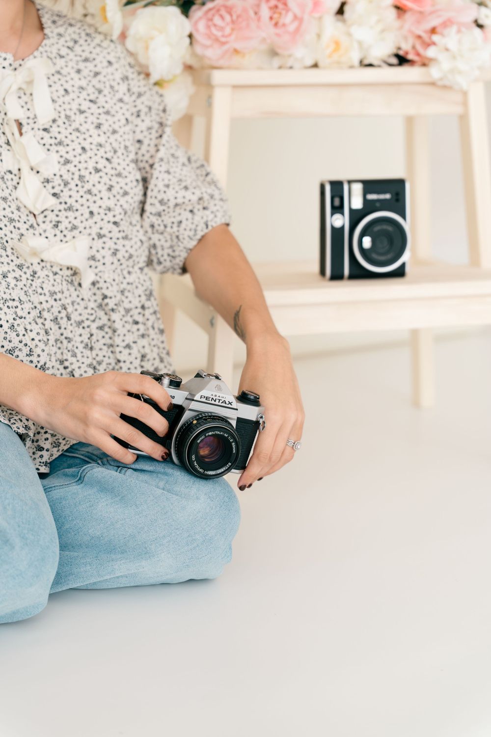 Hands holding vintage camera while wearing floral print top and jeans with pink roses and camera display in background.