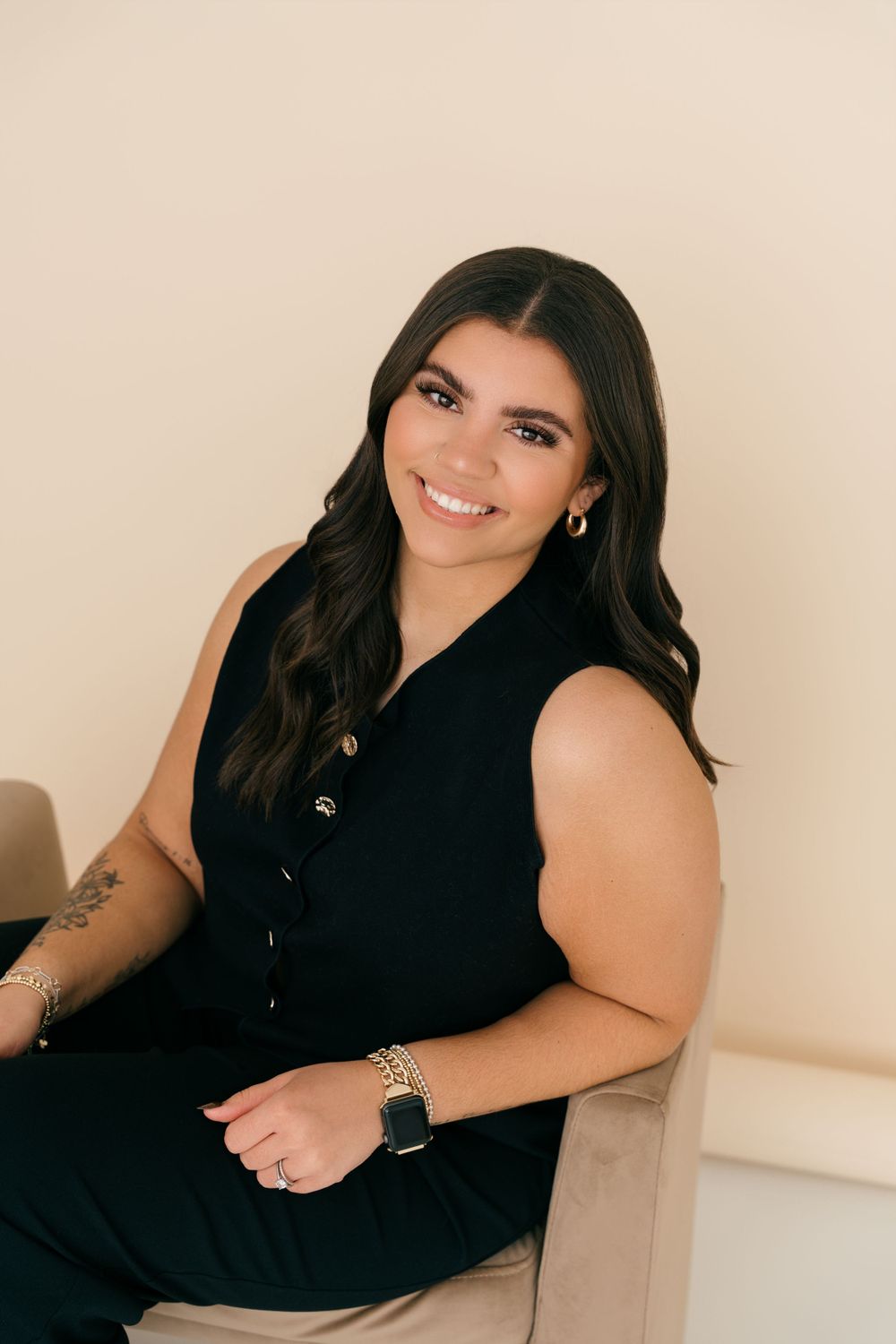 A person wearing a black sleeveless top sits on a beige chair while smiling at the camera in a professional portrait.