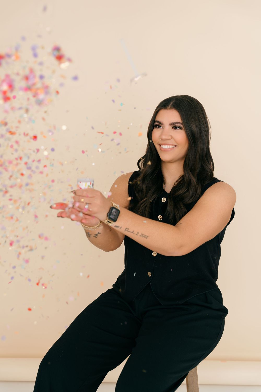 A person in a black outfit celebrates with confetti against a light background while holding a glass.