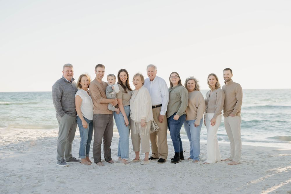 Family group wearing coordinated beige and white outfits standing together on a sunny beach with ocean waves behind them.