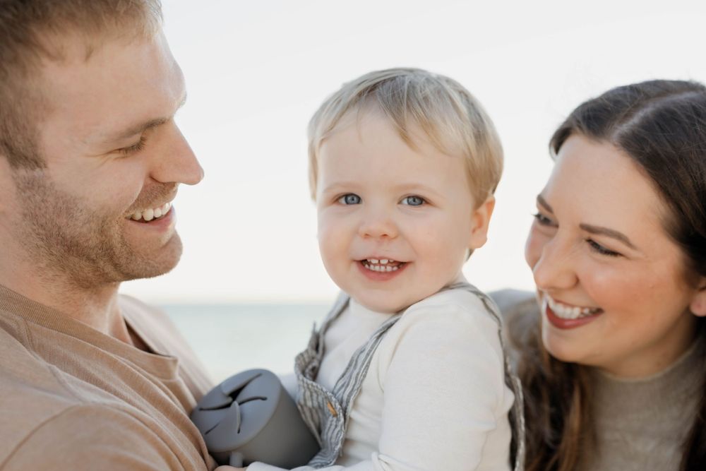 A young child smiles while being held by two affectionate parents in a bright outdoor setting.
