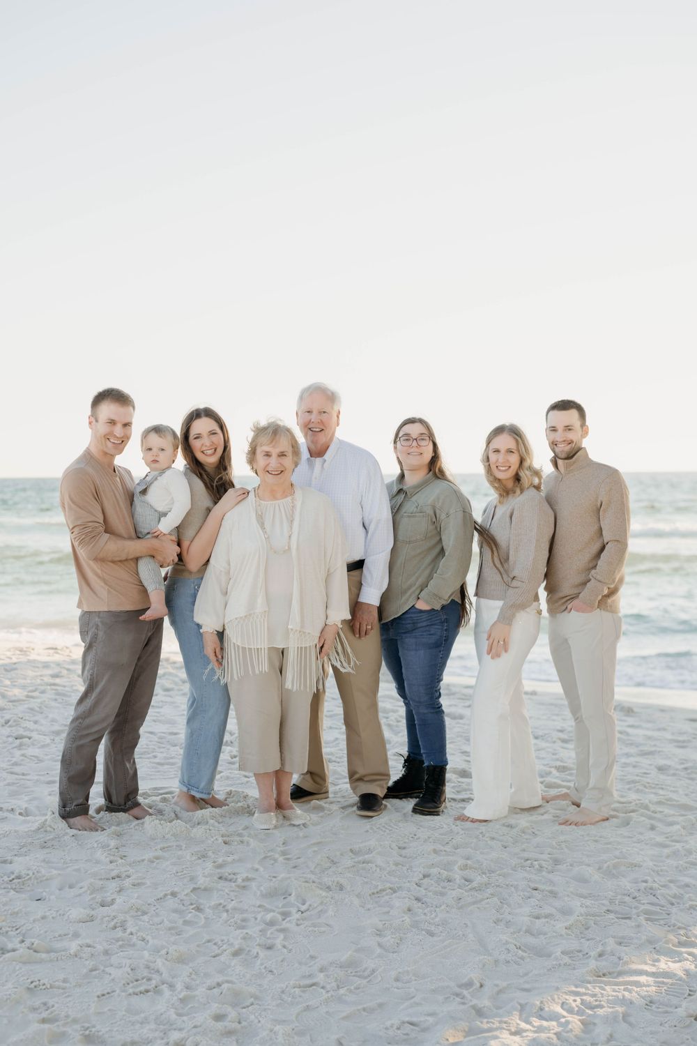 A group of people dressed in neutral colors pose together on a sandy beach at sunset.