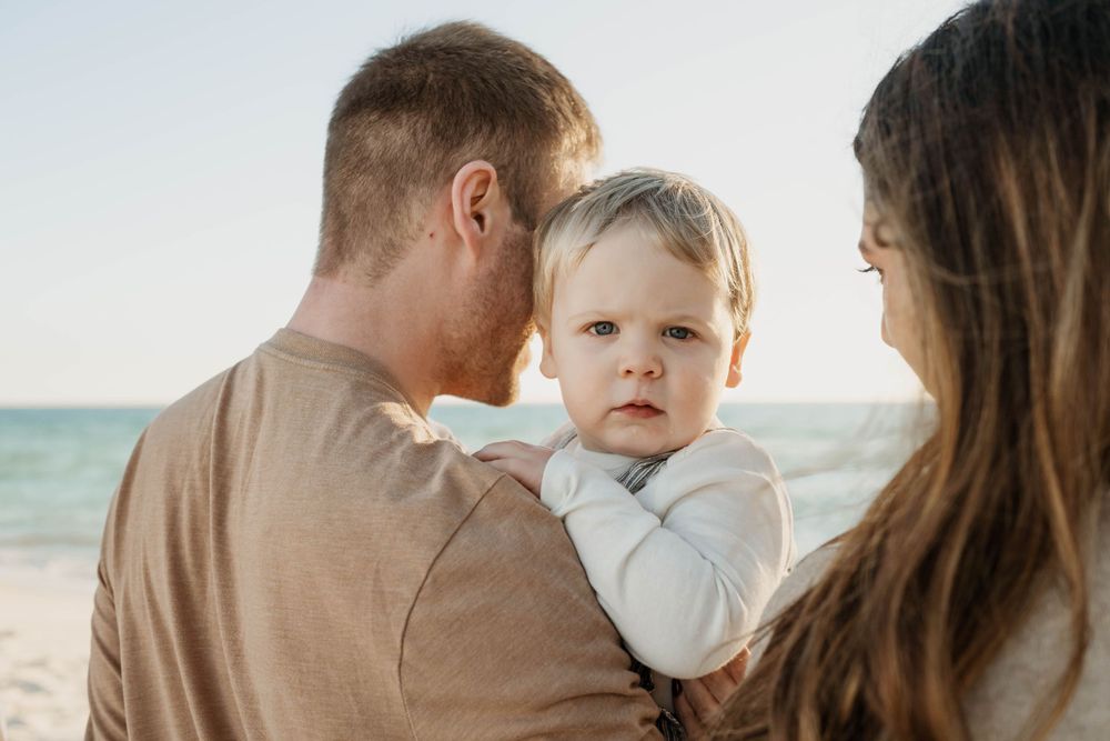 A small family shares a tender moment together on a sunny beach with the ocean in the background.