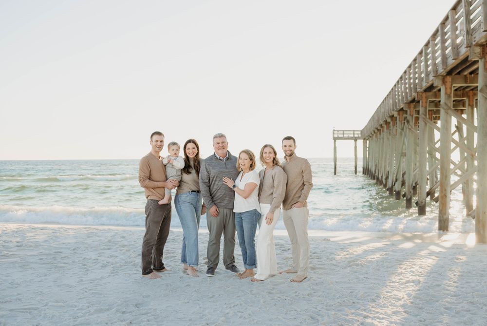 A multi-generational family poses together on a white sandy beach near a wooden pier at sunset.