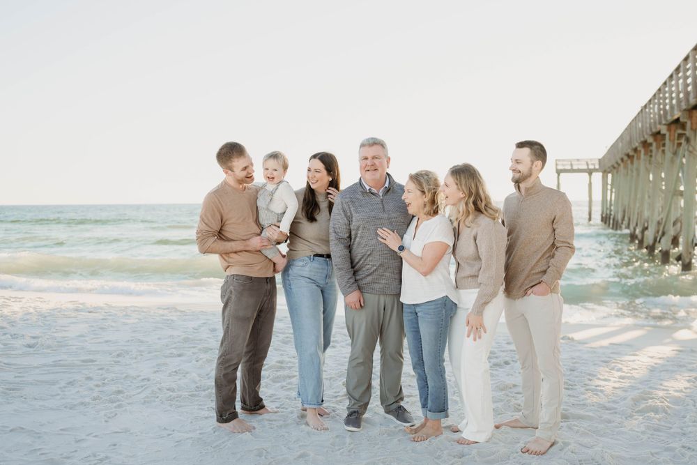 A family group stands together on a sandy beach near a wooden pier during sunset in neutral colored clothing.