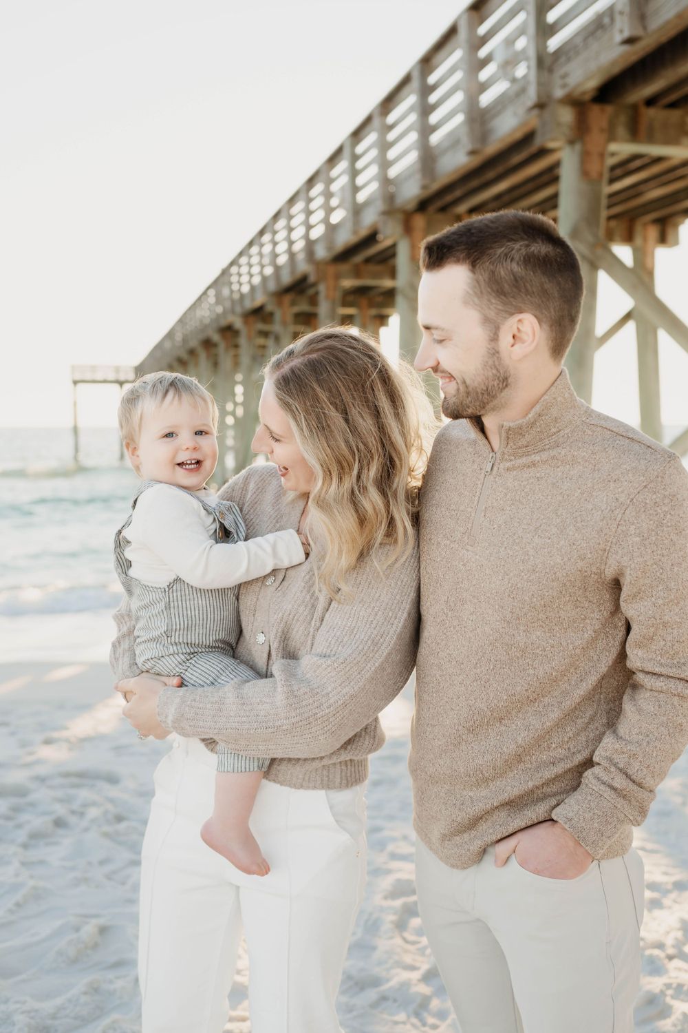 A small family wearing neutral sweaters shares a tender moment on the beach under a wooden pier at sunset.