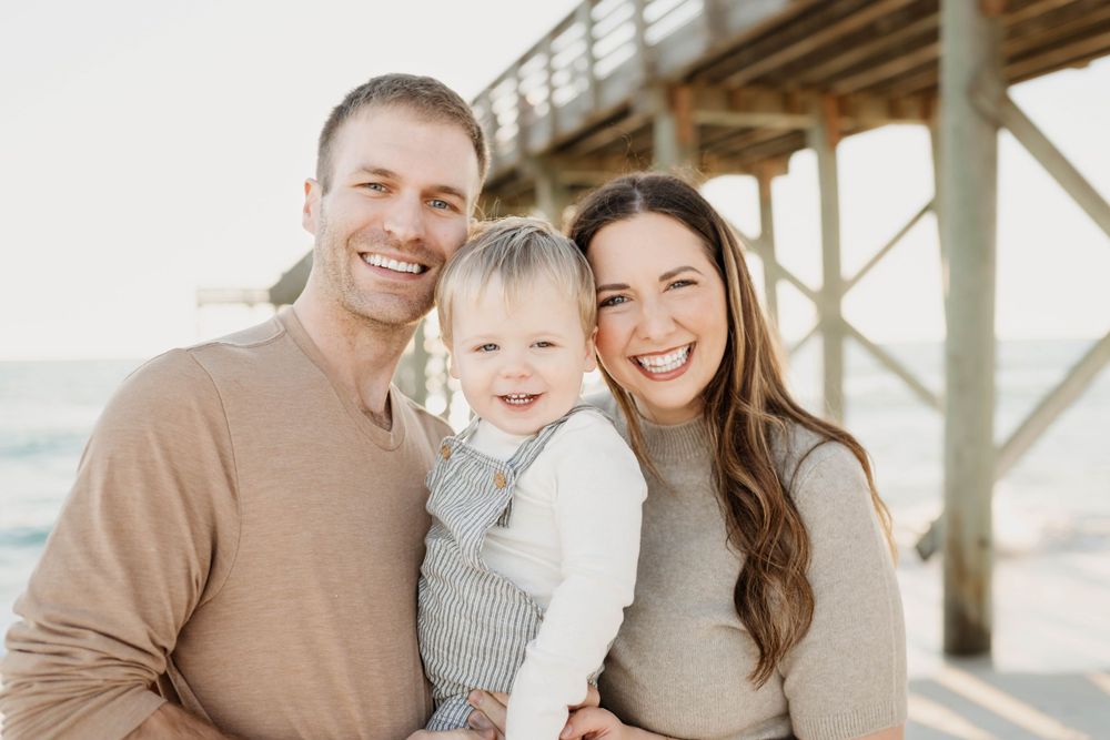 A happy family of three poses together under a wooden pier at the beach on a bright day.