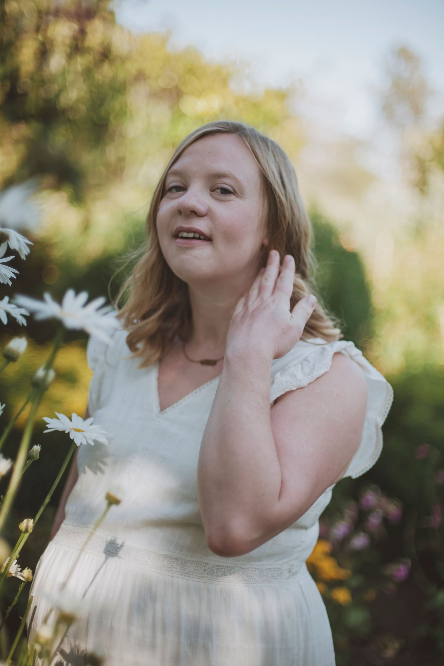 Light and dreamy portrait in a garden with daisies features a pregnant woman in a white dress touching her hair.