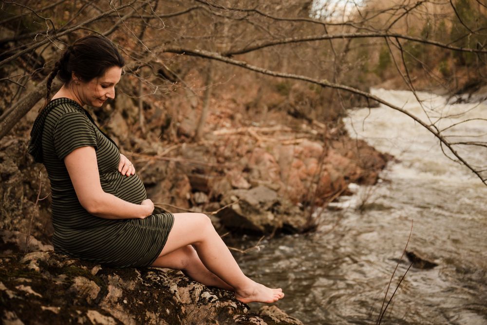 A pregnant woman rests on a fallen log beside a serene stream in autumn.