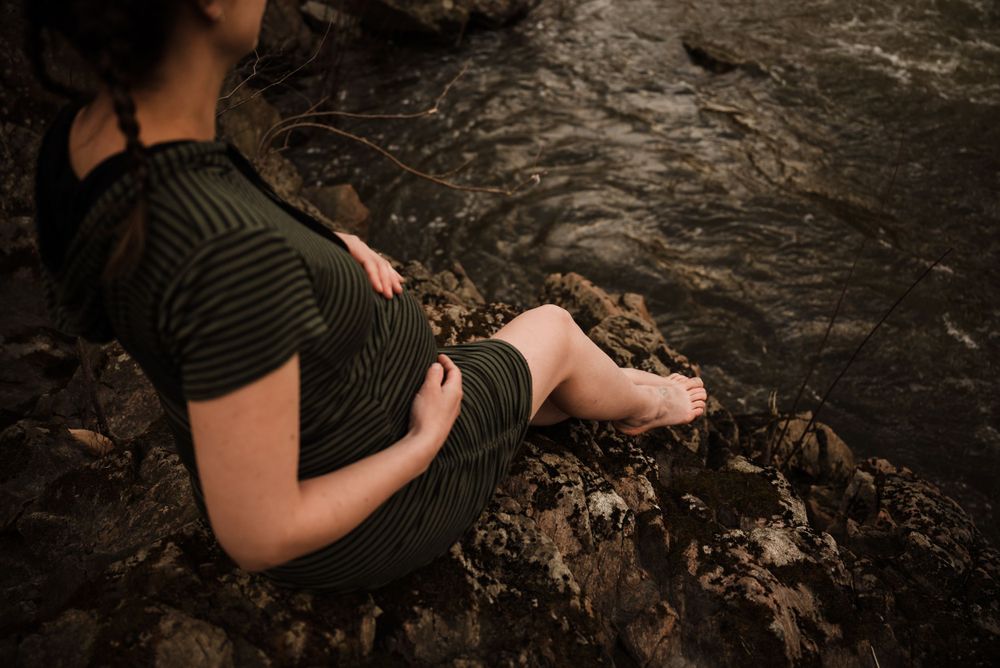 A person in a dark green dress sits by a rocky stream during a maternity photo session.