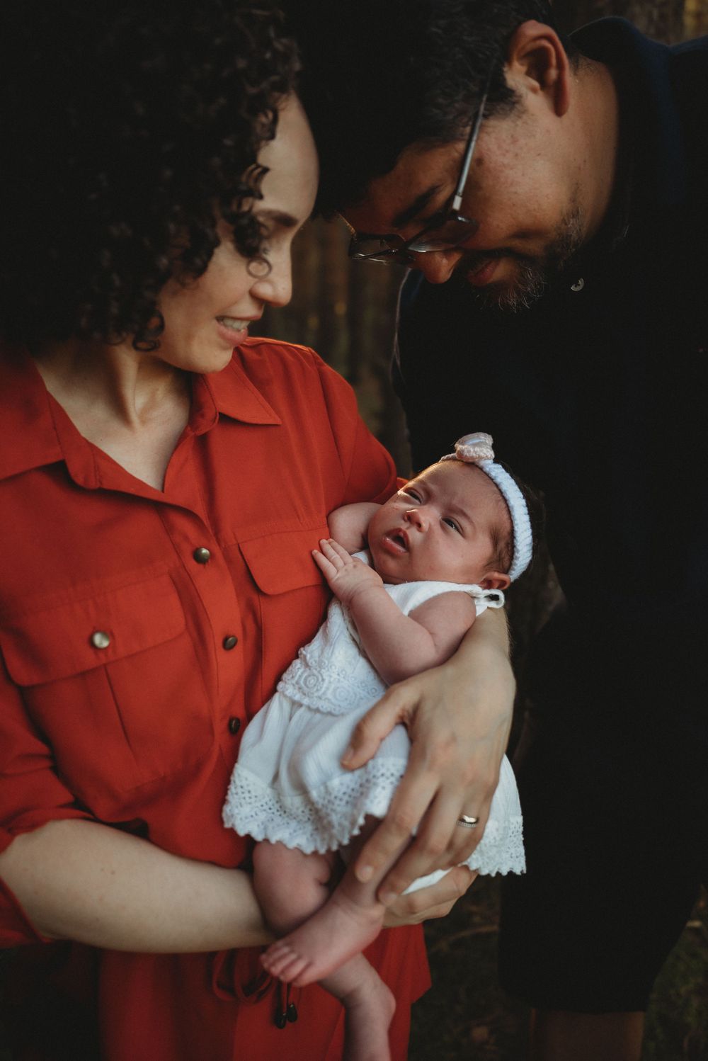 A tender family moment as parents gaze lovingly at their newborn baby wearing a white dress and headband.