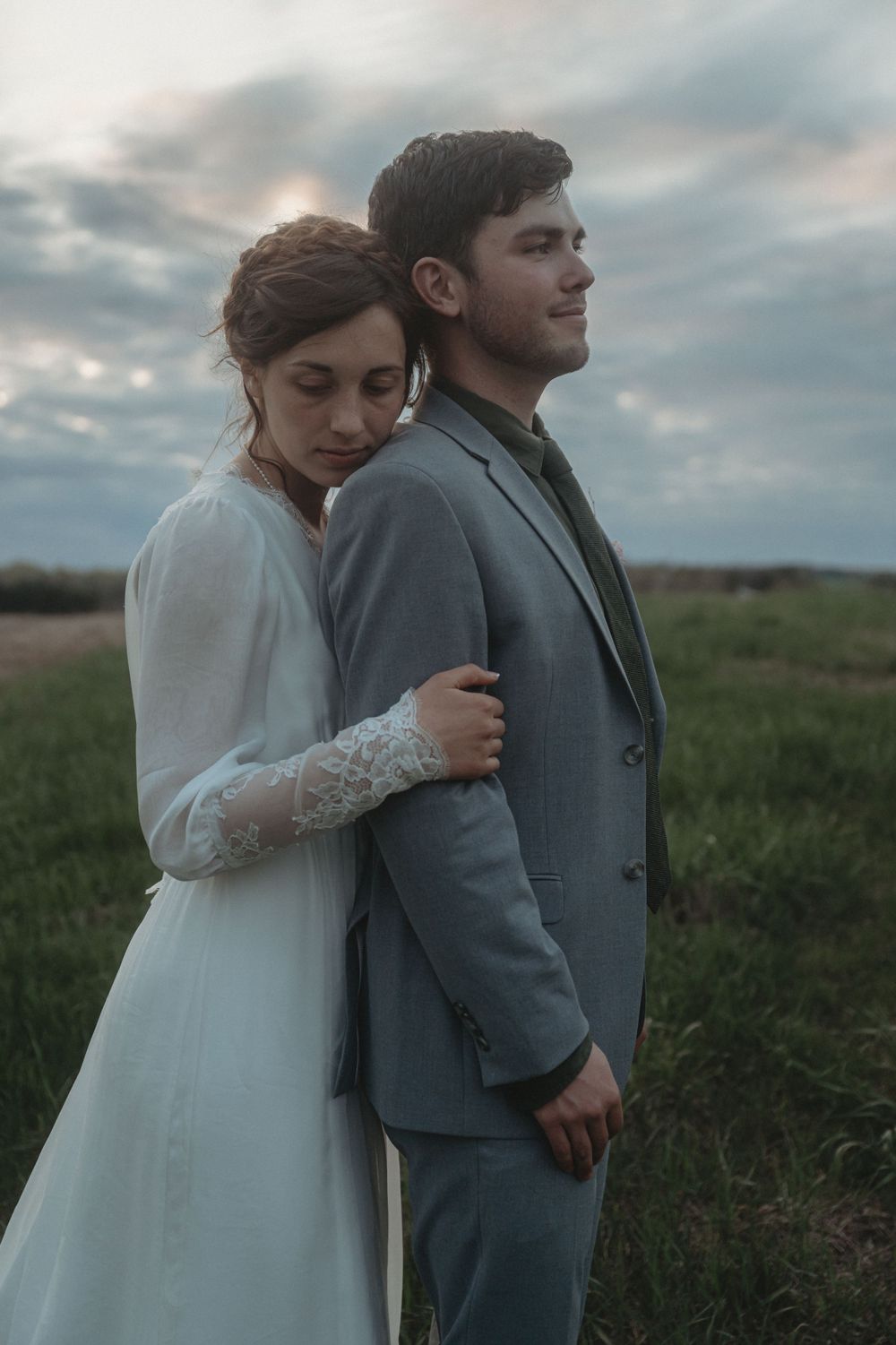 A quiet moment between newlyweds in formal attire standing in a field at dusk.