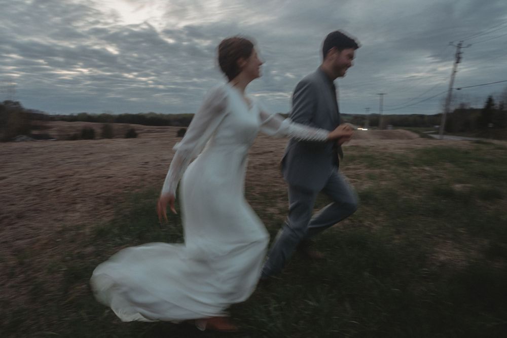 A couple in wedding attire runs playfully through a field at dusk with the bride's dress flowing behind.